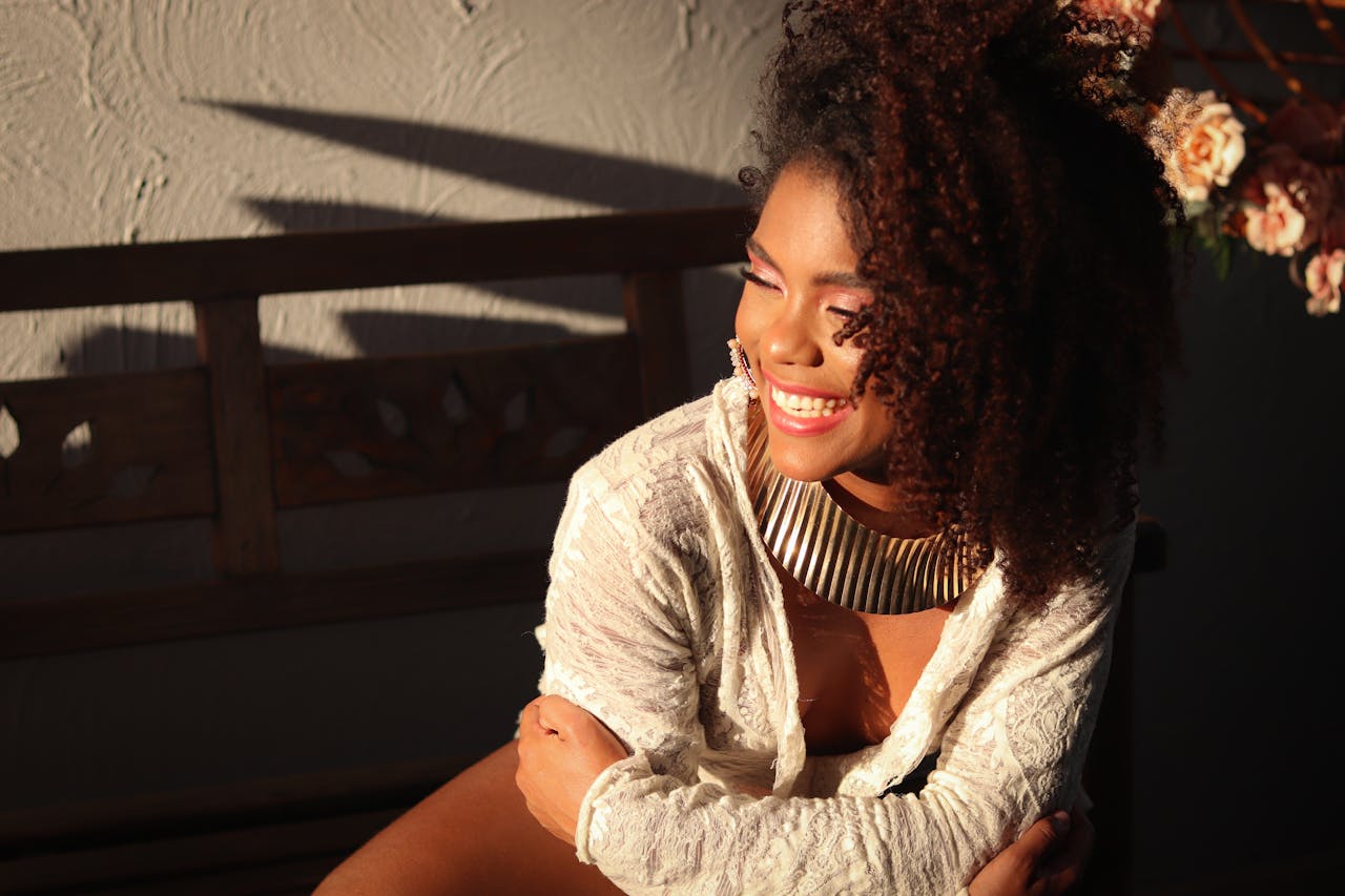 A joyful woman with an afro smiles warmly in a sunlit room, wearing a stylish necklace.